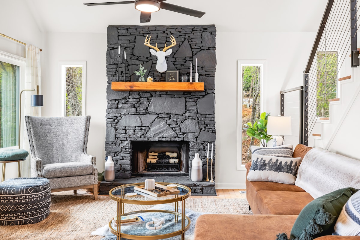 A modern living area is centered around a black stone fireplace, accented with a wooden mantel. Natural light enters through large windows, highlighting a mix of seating options, including a plush brown couch and a textured gray armchair. A circular coffee table holds various decor items.