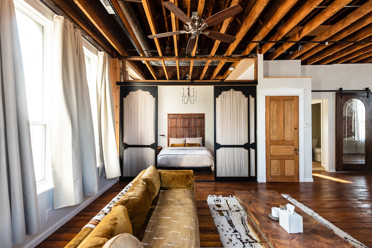 A spacious bedroom features high wooden ceilings with exposed beams and ample natural light from large windows. A comfortable bed is framed by elegant paneling, while a stylish velvet sofa sits nearby. The room's warm wood flooring adds to the inviting ambiance.