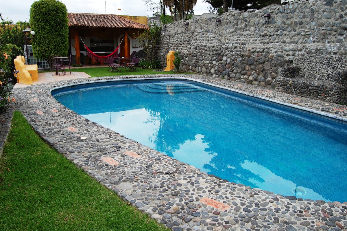 A tranquil swimming pool is framed by natural stone pathways, with a neatly manicured lawn surrounding it. In the background, a shaded seating area is visible, along with a hammock for relaxation. Stone walls provide privacy and a rustic charm to the outdoor space.