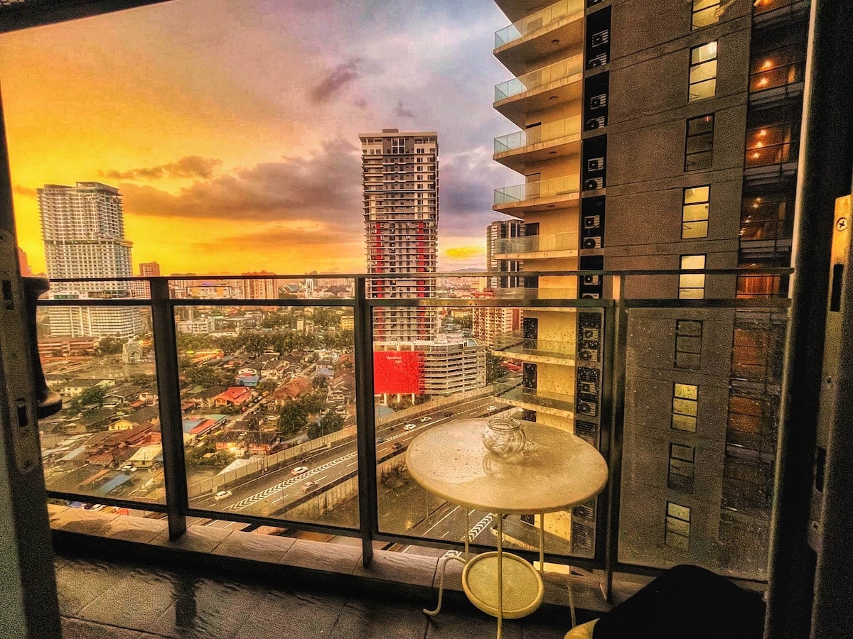 A glass balcony railing frames a stunning urban sunset view, where warm orange and purple hues fill the sky. Below, city streets and buildings create a vibrant skyline, while a small round table with a glass sits on the balcony floor.