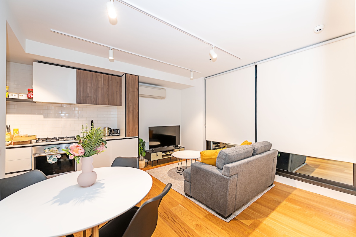 A modern living area features an open layout with a round dining table and four black chairs. A comfortable gray sofa sits across from a wall-mounted television. Natural light is provided by large windows, partially covered with blinds, and the kitchen area is visible in the background.