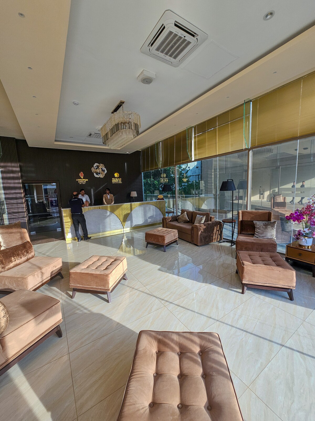 A welcoming lobby area is furnished with several brown lounge chairs and ottomans, arranged around a decorative coffee table. A reception desk is positioned at the back, attended by staff. Large windows allow natural light to brighten the space, highlighting the polished tile flooring.