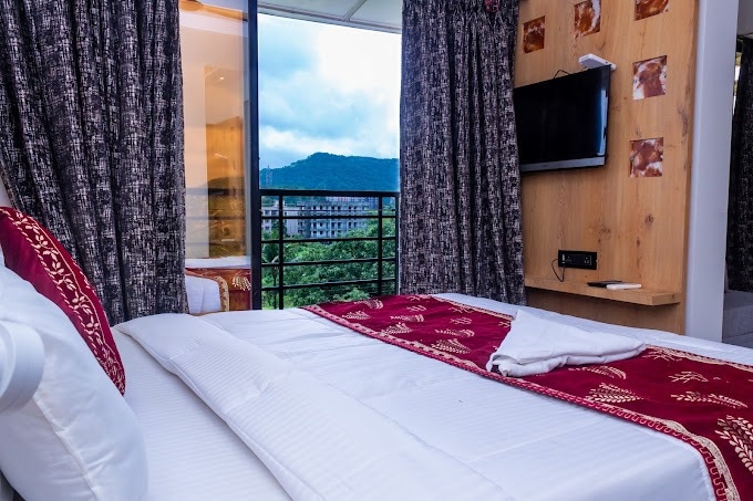 A comfortable sleeping area features a spacious bed dressed in crisp white linens and a deep red decorative throw. Large windows offer views of the surrounding hills, framed by patterned curtains. A wall-mounted television is positioned nearby, enhancing the modern decor.