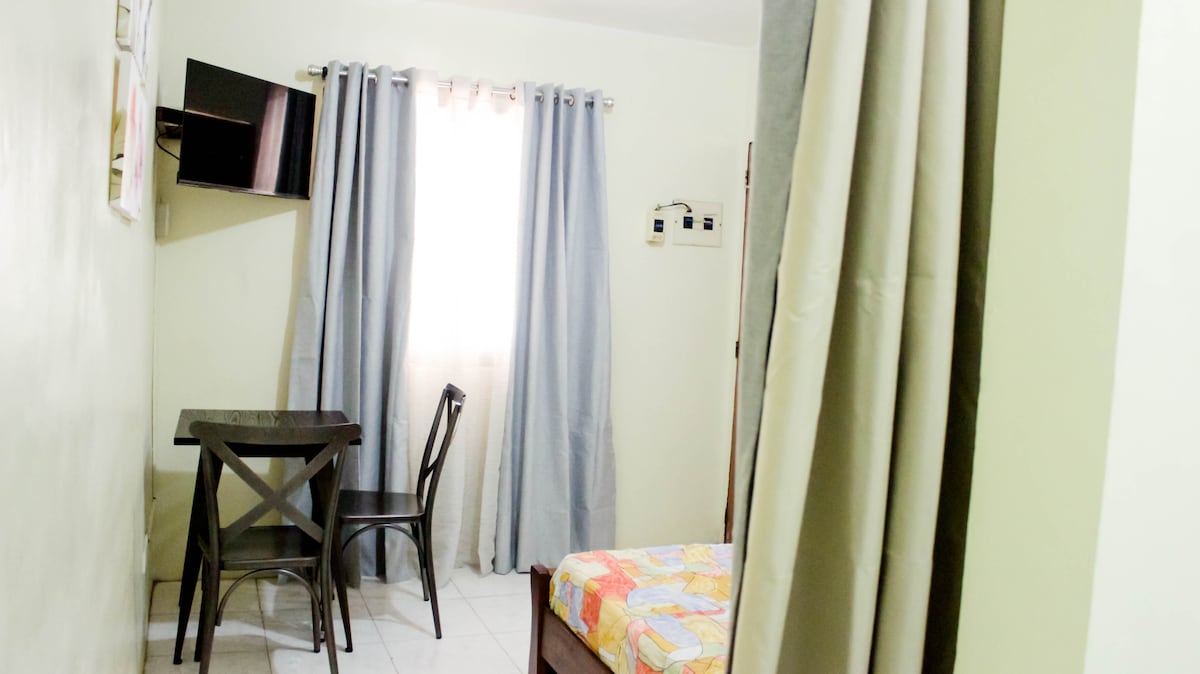 A compact room features a small dining table and chair set near a window dressed with light curtains, allowing natural light to filter in. A colorful quilt adorns the bed, and a flat-screen television is mounted on the wall, enhancing the functionality of the space.