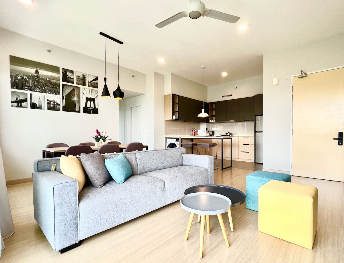 A modern living area is presented with a light grey sofa adorned with decorative cushions in blue and yellow. A dining area is visible behind, featuring a medium-sized table with chairs. The fully equipped kitchen includes contemporary cabinetry and appliances. Decorative wall art adds a subtle touch.