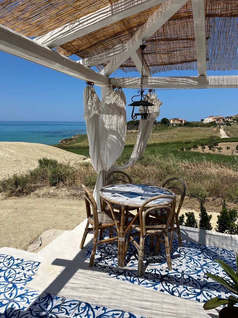 A shaded outdoor seating area features a round table with two rattan chairs set against a backdrop of the Mediterranean Sea. Light-colored fabric drapes gently from the canopy, complementing the blue and white patterned tiles on the ground, while lush greenery borders the scene.