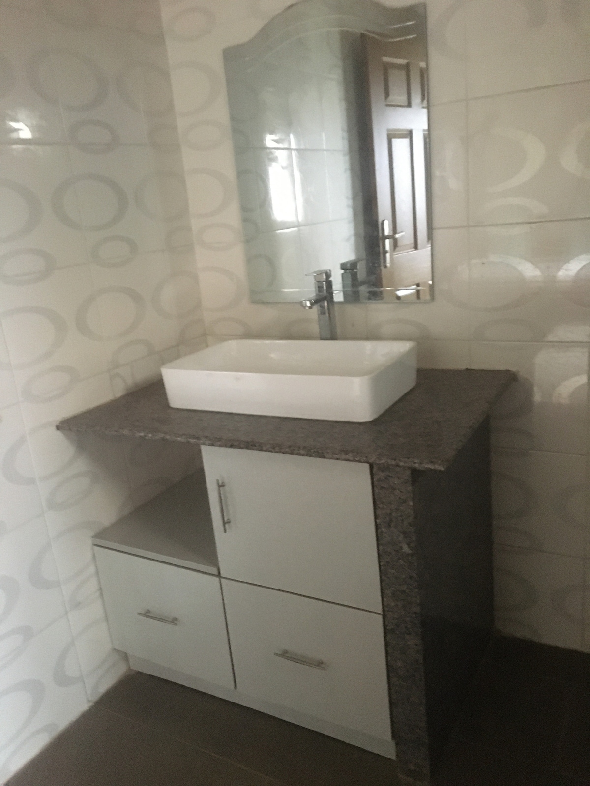 A clean and modern bathroom setup features a sleek, rectangular sink atop a granite countertop. Below the counter, storage is provided by two cabinets with minimalistic hardware. A wall-mounted mirror enhances the space, reflecting the well-lit tiles surrounding the area.