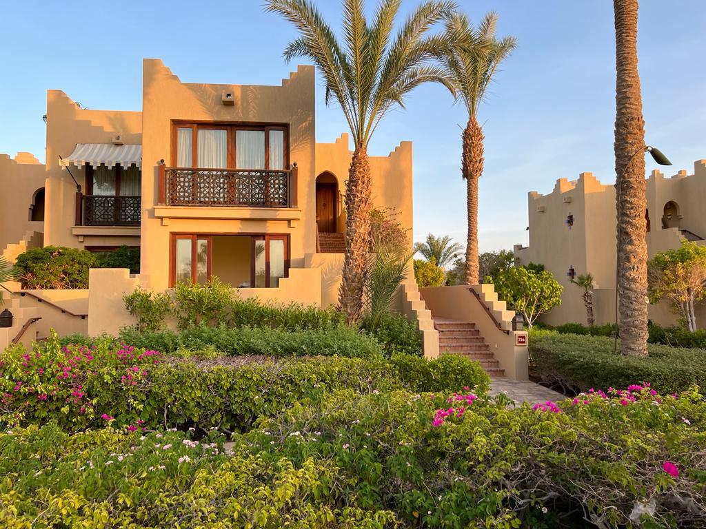 A charming exterior of a two-story building is surrounded by lush greenery and vibrant flowers. Palm trees rise beside the structure, which features a balcony with wooden railing and smooth, beige walls that reflect the warm hues of the setting sun.