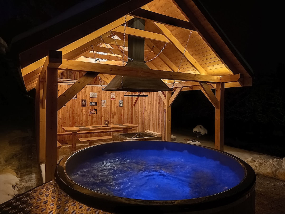 A wooden shelter features an inviting hot tub with blue water illuminated by soft lighting. The structure is supported by timber beams and includes a seating area adorned with wooden benches. Snow can be seen surrounding the space, enhancing the tranquil atmosphere.
