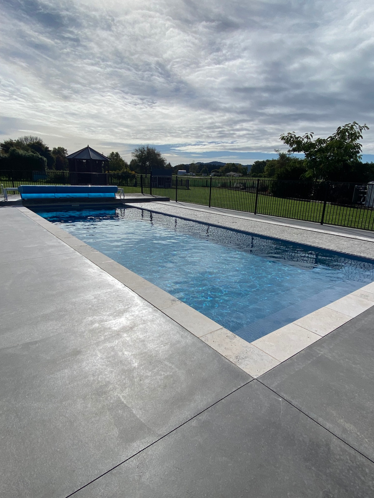 A serene swimming pool is framed by a minimalist paved area and surrounded by a secure black fence. Lush greenery and distant hills are visible beyond the pool, while a shaded gazebo can be seen in the background under an open sky.