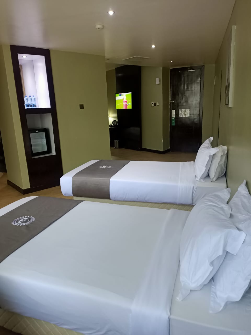 The sleeping area features two spacious beds with light-colored linens and dark accent throws. A small refrigerator is visible in the corner, and a wall-mounted television is mounted across from the beds. Neutral tones dominate the interior, accompanied by soft, ambient lighting.
