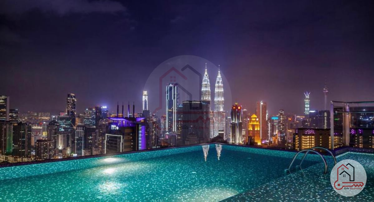 An infinity pool overlooks the city skyline, featuring shimmering water illuminated by nearby lights. Iconic skyscrapers, including the PETRONAS Towers, are visible in the background against a night sky filled with stars.