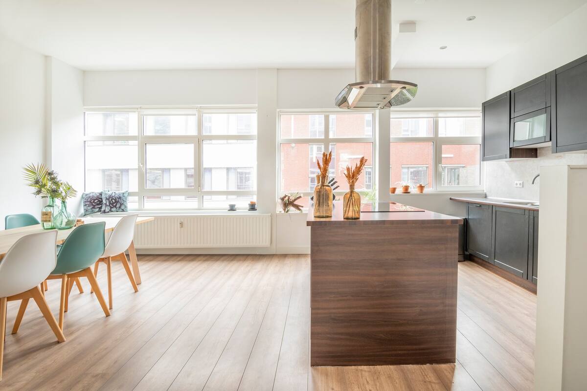 An open kitchen and dining area is displayed, featuring a central island with a range hood. A dining table with colorful chairs accommodates multiple guests, while natural light filters through large windows, highlighting the wooden flooring and decorative elements placed throughout the space.