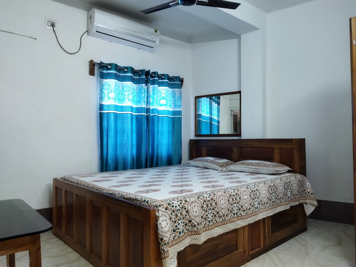 A spacious bed is positioned against a light-colored wall, adorned with a patterned bedspread. Dark curtains with a blue design frame a nearby window, while a ceiling fan provides ventilation. A wooden table stands beside the bed, enhancing the room's functionality.