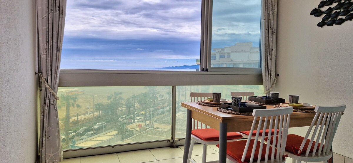 A dining table with white chairs and a wood finish is set near a window, offering a view of the sea and coastal landscape. Natural light fills the space, highlighting table settings that consist of dishware and utensils, creating a warm atmosphere for dining.