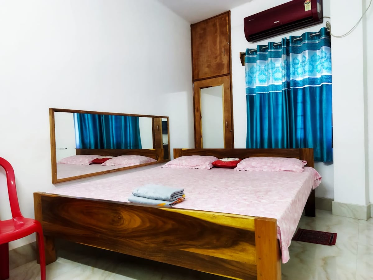 A spacious sleeping area features a large wooden bed with pink bedding, complemented by a matching set of pillows. A framed mirror captures the room's light, while blue curtains adorn the window above. A red chair adds a pop of color to the inviting space.