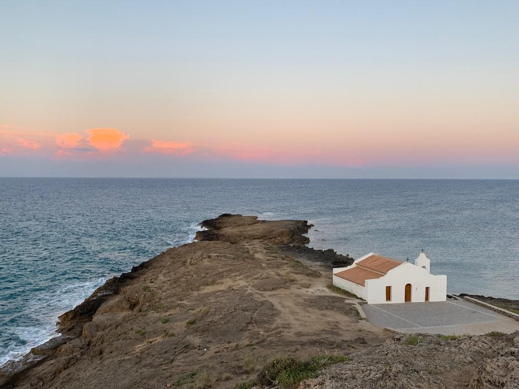 A coastal view showcases a small white chapel set against the expansive sea. The rocky shoreline extends into the water, while the horizon displays soft hues of orange and pink as the day transitions into evening. Gentle waves lap against the rocks.
