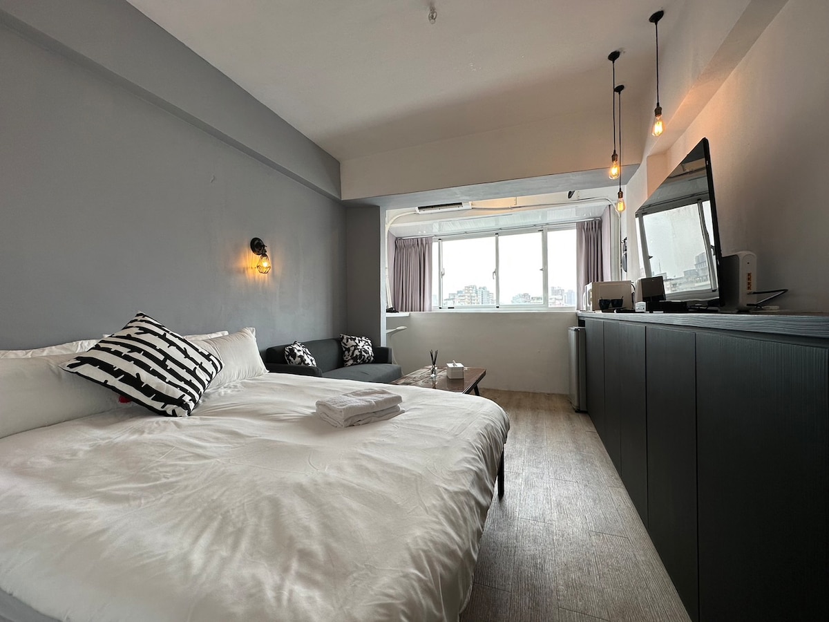 A spacious room features a large bed with crisp white linens, complemented by patterned throw pillows. A cozy seating area is visible beside a large window, allowing natural light to fill the space. A flat-screen TV is positioned atop a dark cabinet against the wall.