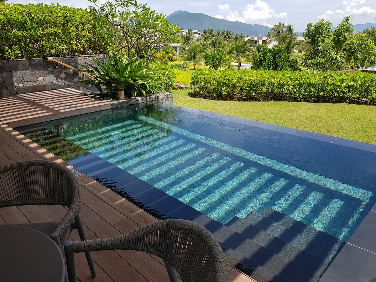 A private pool is framed by natural stone and lush greenery, reflecting the surrounding landscape. Sunlight casts intriguing patterns on the water, while a wooden deck with seating facilitates relaxation and enjoyment of the scenic views.