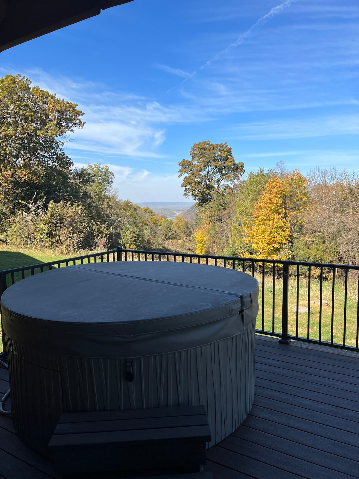 A spacious deck is featured, hosting a covered hot tub with a protective lid. Lush greenery and colorful trees create a serene backdrop against a clear blue sky, offering scenic views of the surrounding landscape.