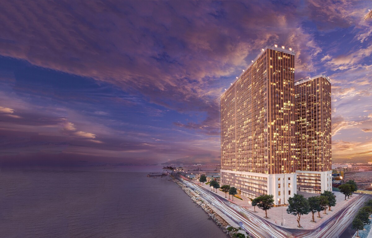 A modern hotel structure rises against a dramatic sky, with hues of orange and purple reflecting off its golden facade. The building is situated near the water's edge, surrounded by a landscaped area and a pathway that follows the shoreline.