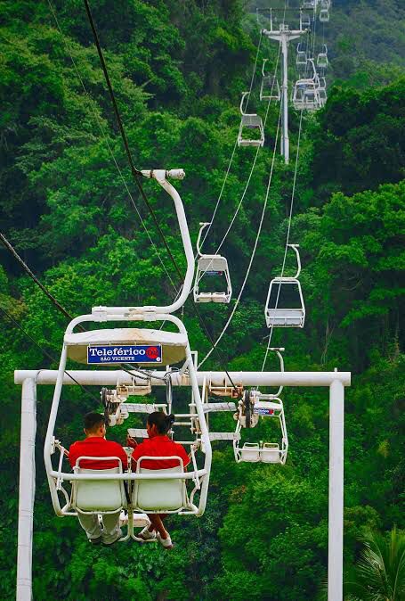 A suspended cable car travels above a lush green landscape, showcasing dense foliage below. Two passengers are seated, enjoying the scenic view. The cable car features a white frame and carries multiple hanging seats along the route.