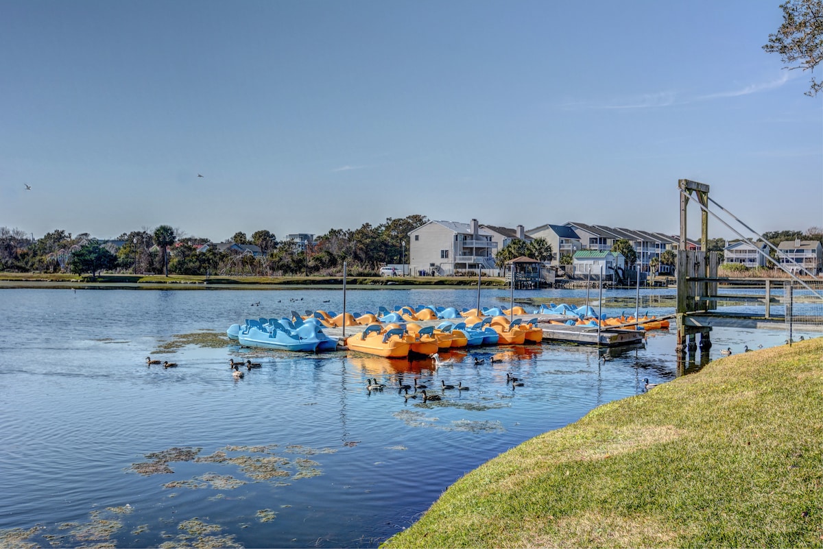 A serene waterway is framed by green grass, featuring colorful paddle boats neatly arranged along a dock. Ducks are seen swimming in the calm water, while trees and buildings create a peaceful backdrop.