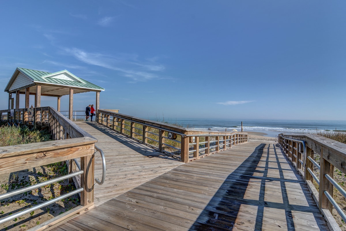 A wooden boardwalk leads from a beach access point towards the ocean. The structure has a white pavilion with a green metal roof in the background. Gentle waves can be seen in the distance, under a clear blue sky.