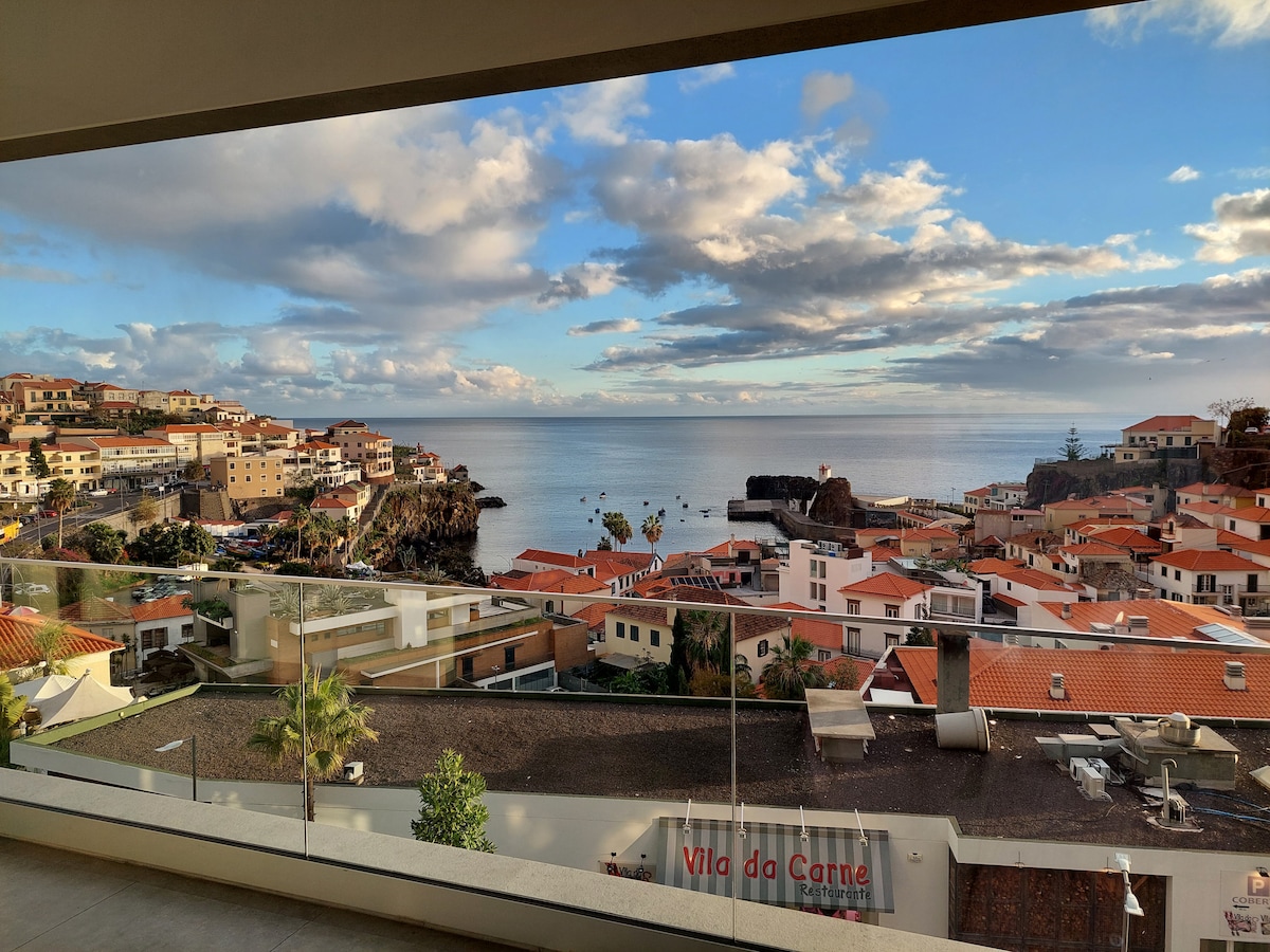 A panoramic view is presented from the balcony, showcasing the coastal landscape of Câmara de Lobos. The scene captures the ocean's horizon where it meets the sky, along with terracotta rooftops and the vibrant greenery of the surrounding area.