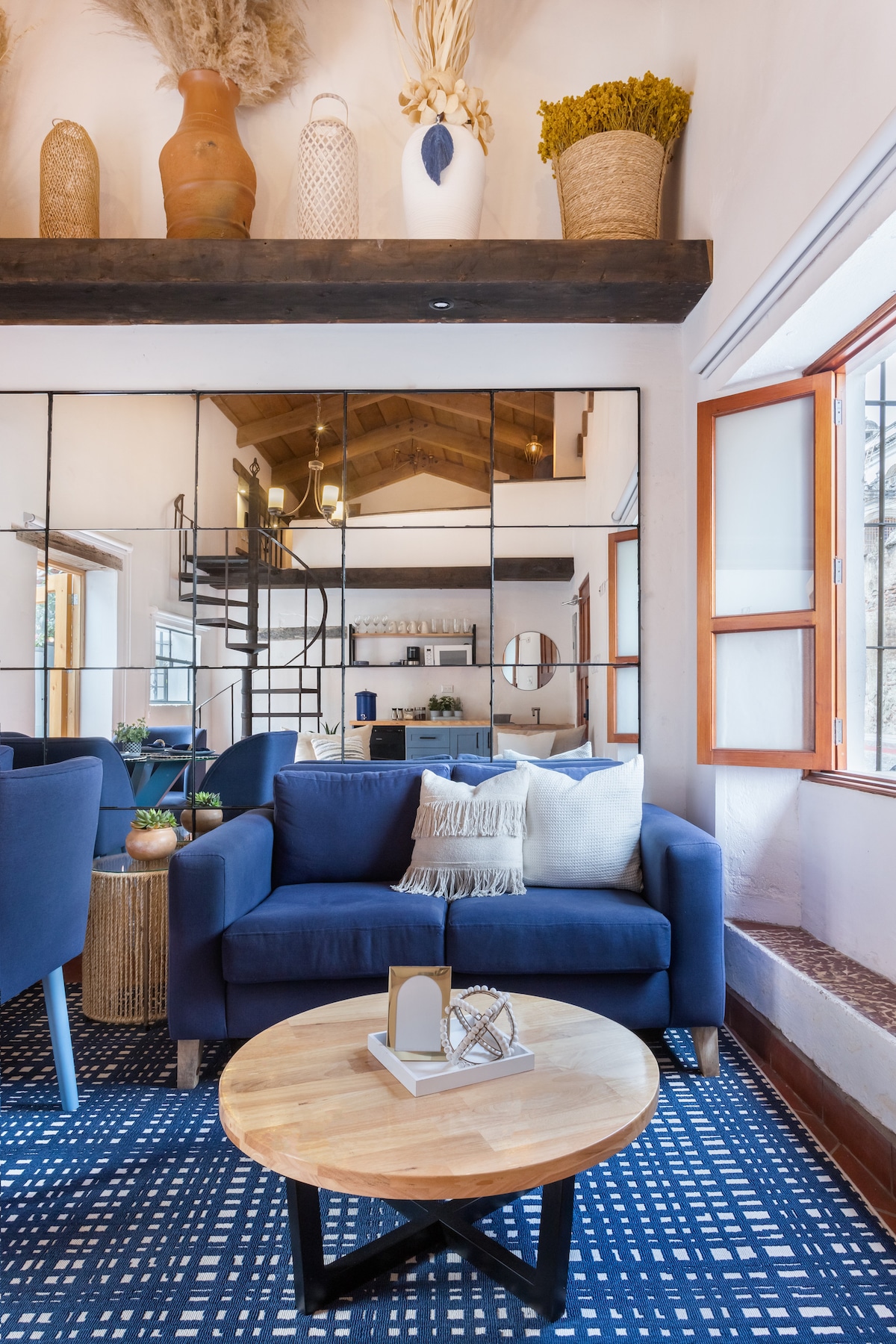 A cozy seating area is framed by large mirrors reflecting the light. A navy blue sofa, adorned with decorative pillows, faces a round wooden coffee table. Potted plants and decorative elements are arranged on a shelf above, enhancing the space's inviting feel.