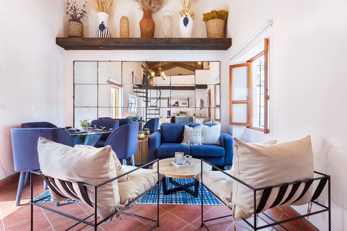 A spacious common area features comfortable seating with soft cushions arranged around a low coffee table. Natural light enters through two windows, illuminating the neutral color palette and highlighting the modern decor that includes a mix of textures and materials.