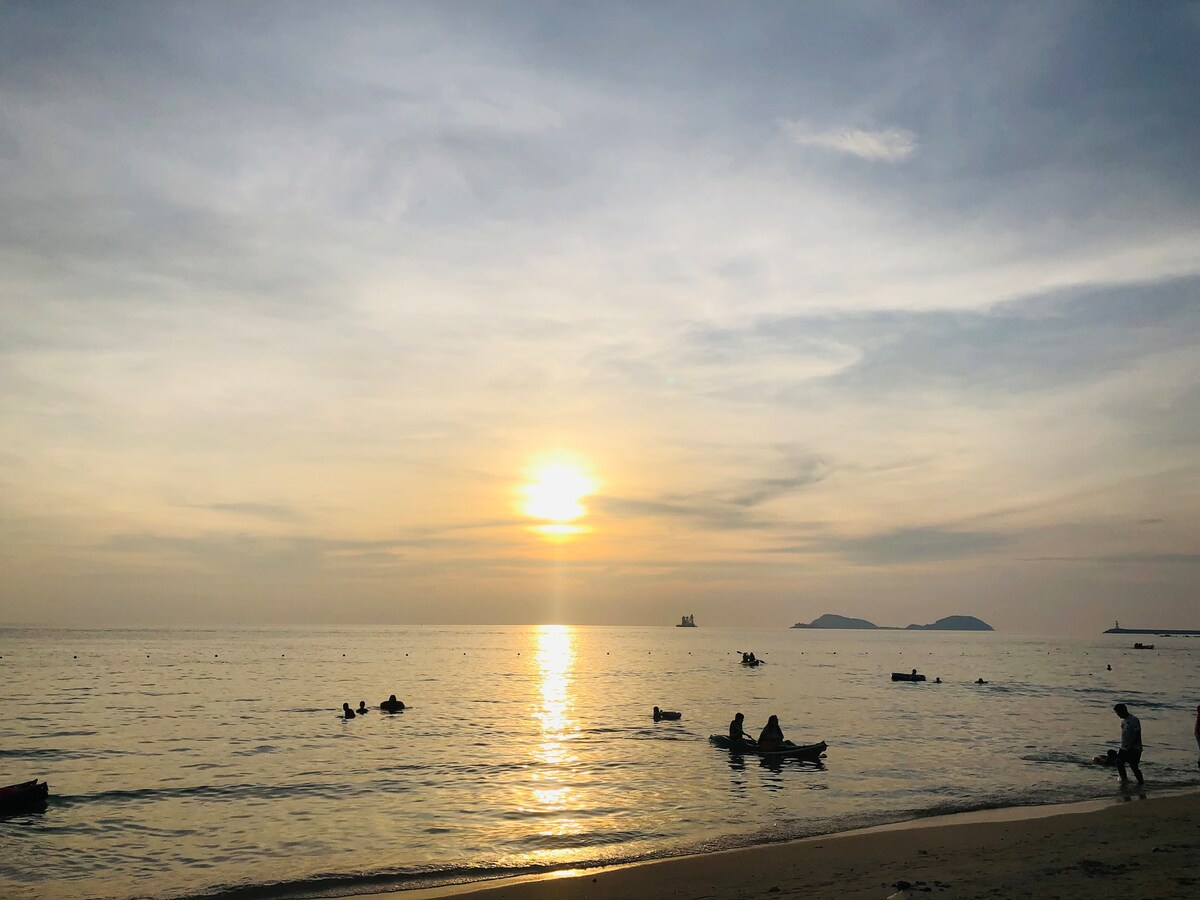 The image captures a serene beach scene at sunset, where gentle waves reflect golden hues. Silhouettes of kayakers can be seen in the water, with distant islands visible across the horizon. The soft clouds in the sky create a tranquil atmosphere.
