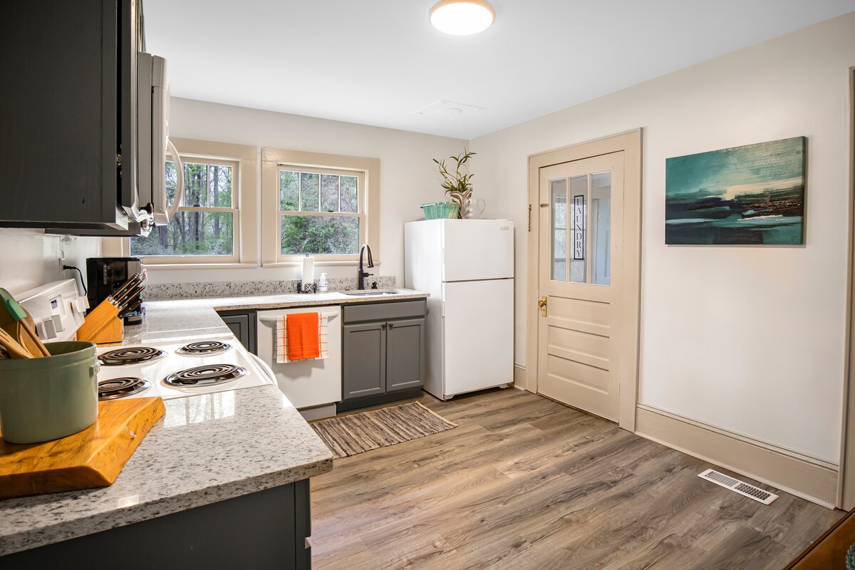 A well-equipped kitchen is displayed, featuring dark cabinetry and a granite countertop. A white refrigerator is positioned next to a door leading outside. Natural light filters through two windows, enhancing the inviting atmosphere. Cookware and utensils are neatly arranged on the countertop.