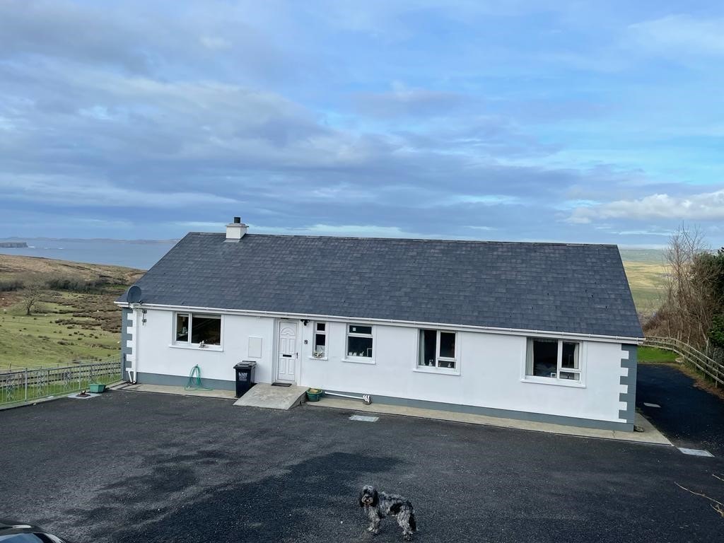 A single-story bungalow is shown, featuring a sloped roof and multiple windows. A small gray dog is visible in the foreground. The driveway is paved, and the surrounding area includes grassy landscapes and distant hills under a blue sky with scattered clouds.