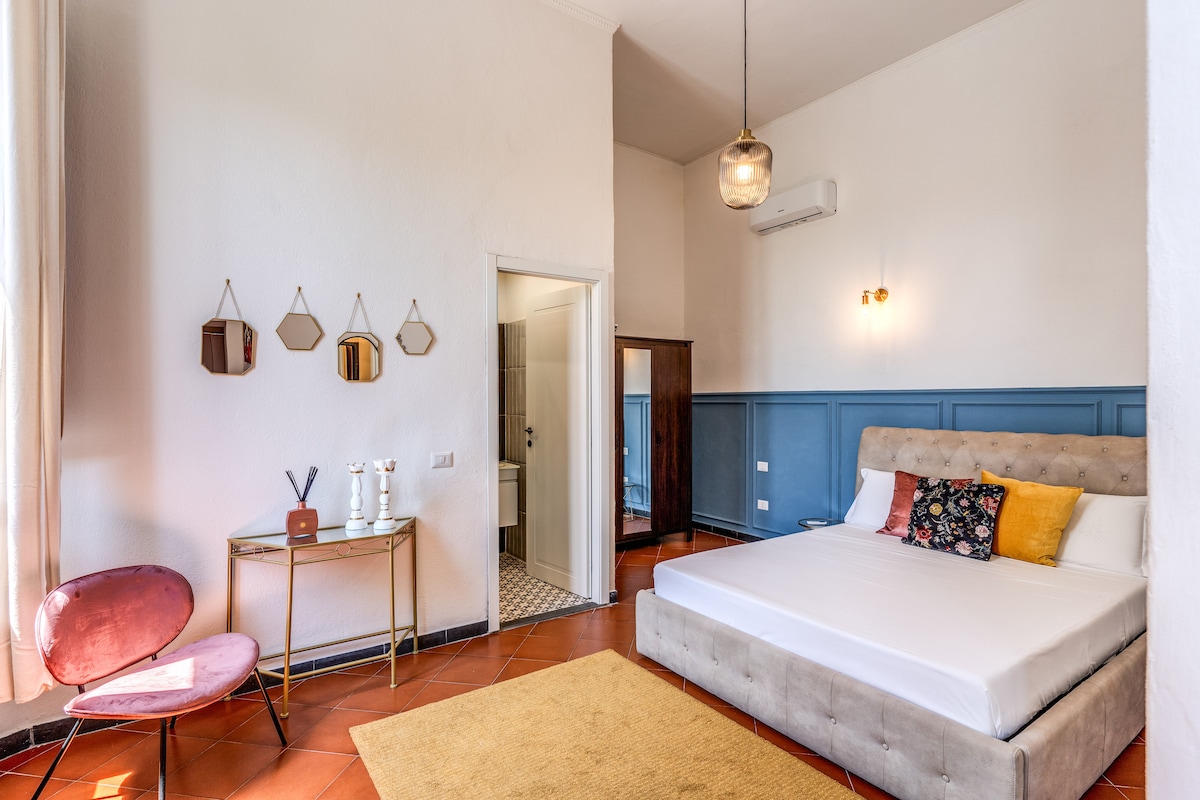 A modern guest room is highlighted by a spacious bed with soft linens and decorative pillows. A small seating area is provided with a pink chair and a gold side table. A doorway leads to an attached private bathroom, and light floods in through a window covered with sheer curtains.