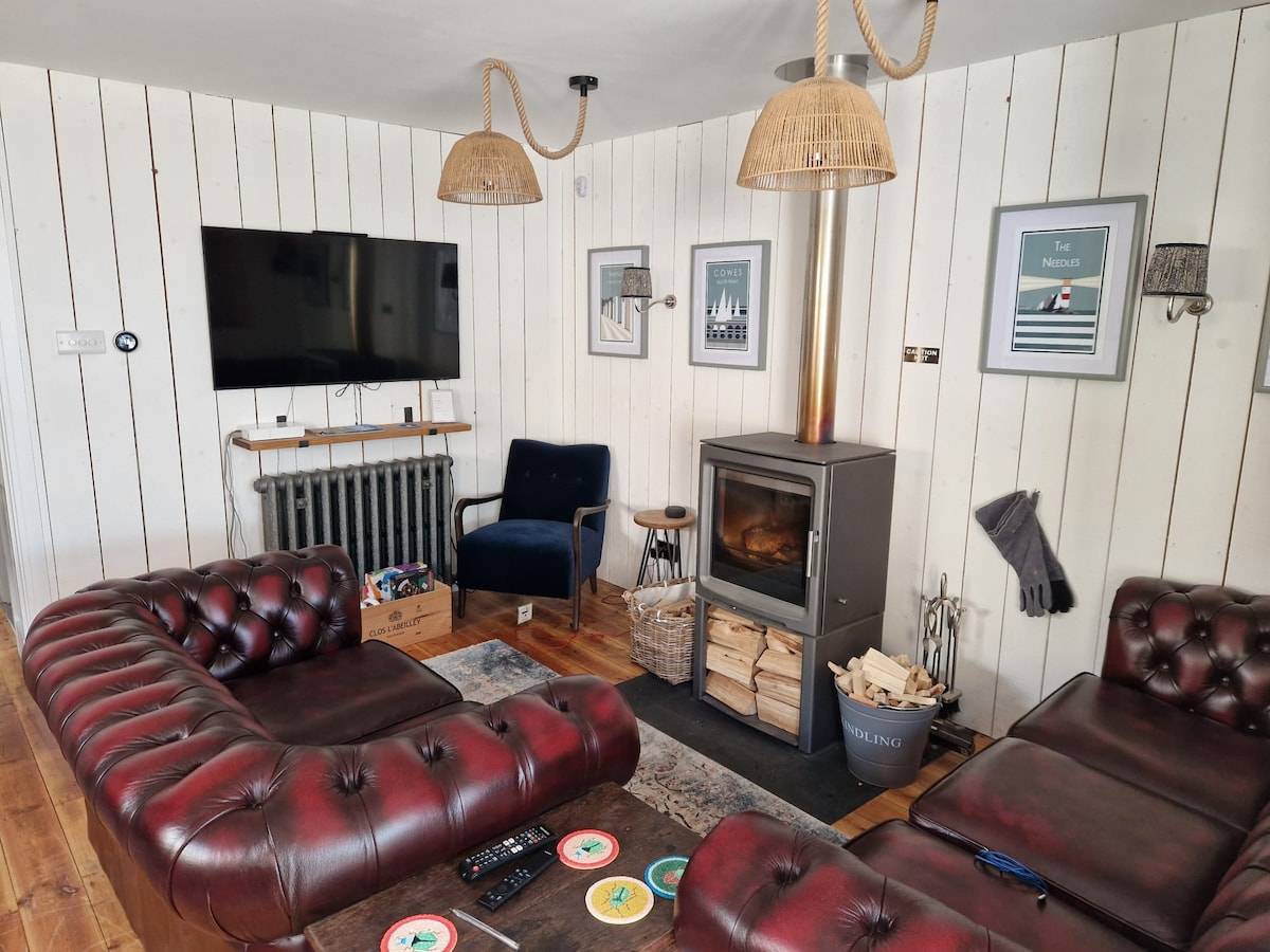 A cozy living space features a log burner as the focal point, surrounded by two leather sofas. A flat-screen television is mounted on a white shiplap wall, with decorative artwork displayed nearby. A small side table holds coasters, and warm lighting is provided by pendant fixtures.