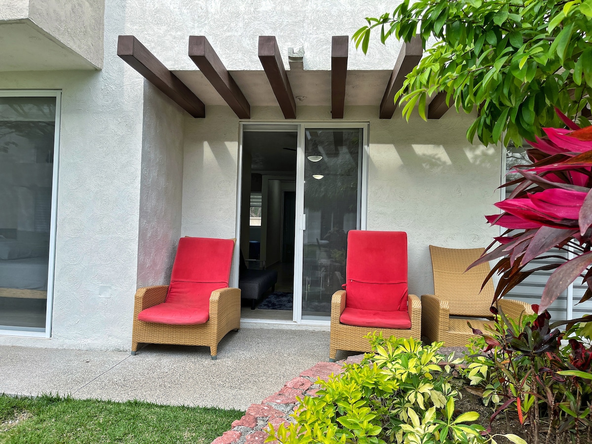 An outdoor seating area features two red lounge chairs placed beside a sliding door. A selection of vibrant plants and greenery surrounds the space, providing a natural touch. The exterior wall is complemented by a wooden beam structure above the chairs.