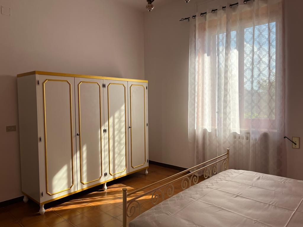 A bright bedroom is filled with natural light streaming through a large window, which is dressed with sheer white curtains. A spacious bed is positioned next to an ornate wardrobe featuring golden accents, providing ample storage space.