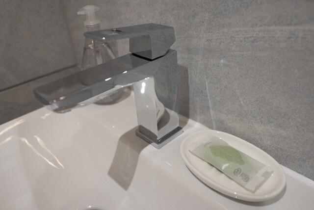 A modern bathroom sink features a sleek chrome faucet, positioned next to a small oval dish holding a single green soap. A clear bottle, likely for liquid soap, is visible in the background against a textured gray wall.