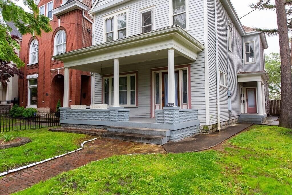 The exterior of the home features a large porch with two prominent columns supporting the roof, surrounded by well-maintained greenery. A paved walkway leads to the entrance, while neighboring homes can be seen adjacent to the property.