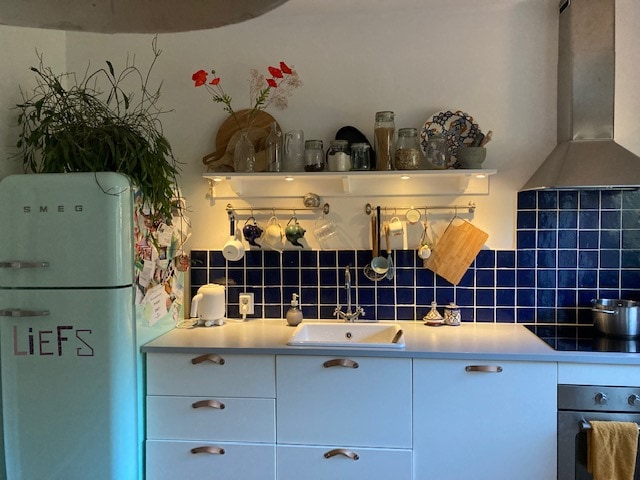 The kitchen features a vibrant blue tiled backsplash, complemented by white cabinetry and brass hardware. A green SMEG refrigerator is prominently displayed. Various kitchenware, including plates and glasses, are arranged on shelves above a white countertop with a sink, creating a functional and inviting cooking space.