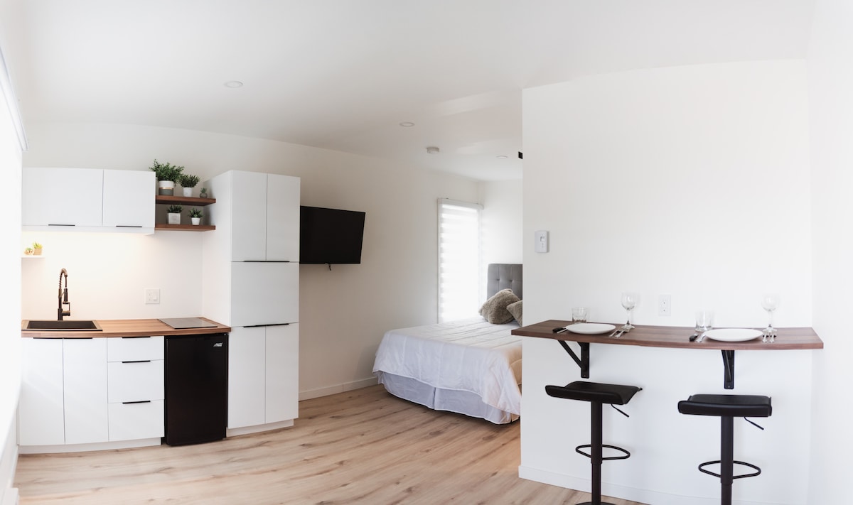 A minimalist studio layout features a kitchenette with sleek white cabinetry and a black refrigerator. A wall-mounted television is visible nearby. Adjacent to the kitchen, a spacious bed is partially dressed in white linens, complemented by a small dining area with two stools set for meals.