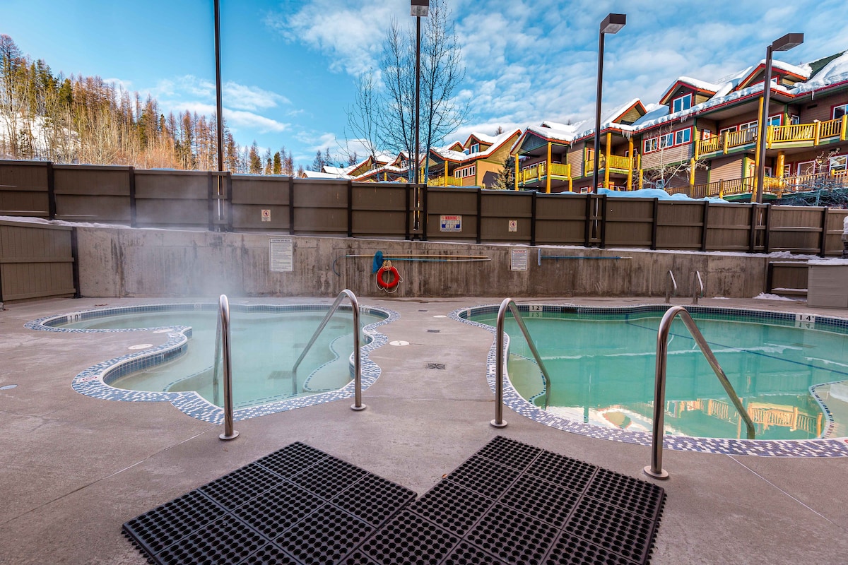 A shared outdoor hot tub and swimming pool are visible, surrounded by a concrete deck. Steam rises gently from the hot tub, while the pool reflects the blue sky and surrounding buildings. Several light posts provide illumination, enhancing the relaxing outdoor atmosphere.