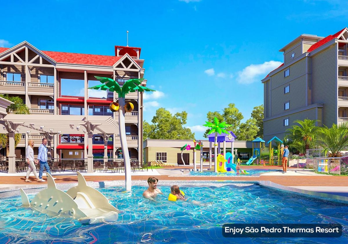A family-friendly outdoor pool area features palm tree accents and playful water attractions. Colorful slides and water play structures are visible in a spacious, shallow section of the pool. Surrounding the area, lounge spaces and resort buildings create a lively atmosphere.