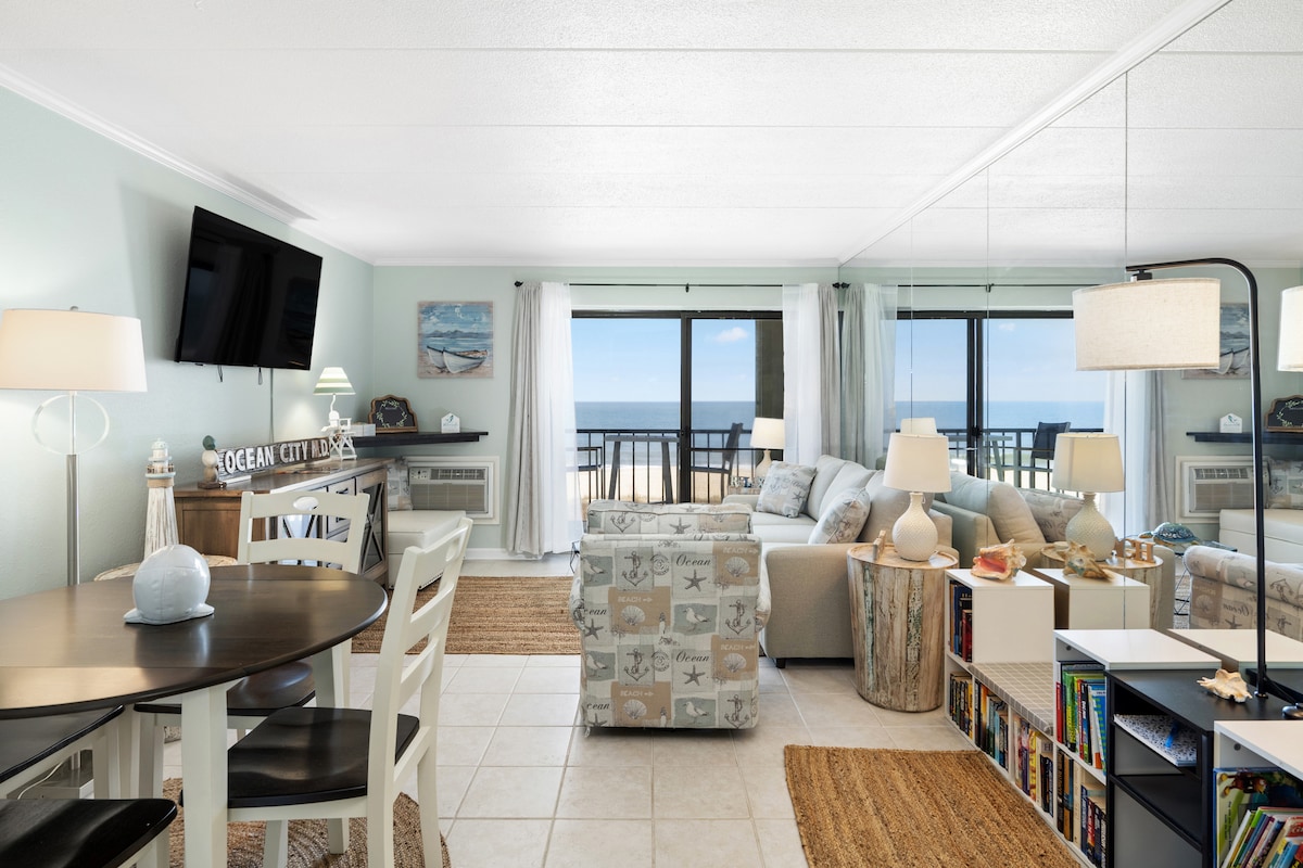 A bright living area features large windows that overlook the ocean, with comfortable seating arrangements. A small dining table for four is paired with white chairs. Light-colored walls create an airy feel, while a colorful bookshelf adds a pop of interest to the space.