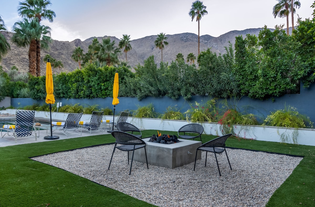 A fire pit is surrounded by four black chairs, set within a gravel area. Yellow umbrellas provide shade nearby, while lounge chairs are positioned for relaxation. Lush greenery and mountainous terrain create a serene backdrop in this outdoor space.