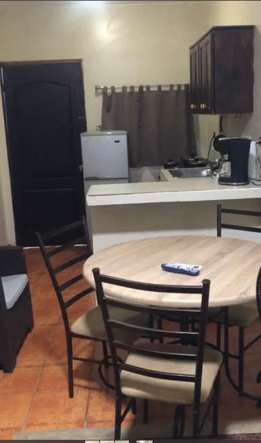 A compact kitchen area features a round wooden dining table with four chairs. Dark cabinetry is visible above a countertop, with a small refrigerator and coffee maker positioned nearby. A window covered by a curtain allows natural light into the space.
