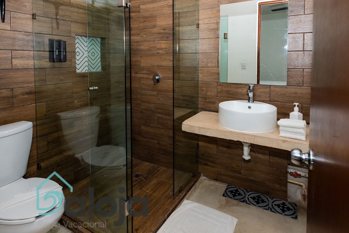 A modern bathroom features a spacious walk-in shower with glass doors and a sleek stone countertop sink. Neutral wood-paneled walls complement the contemporary design, while white towels are neatly arranged beside the sink. A decorative tile pattern adds interest to the floor.