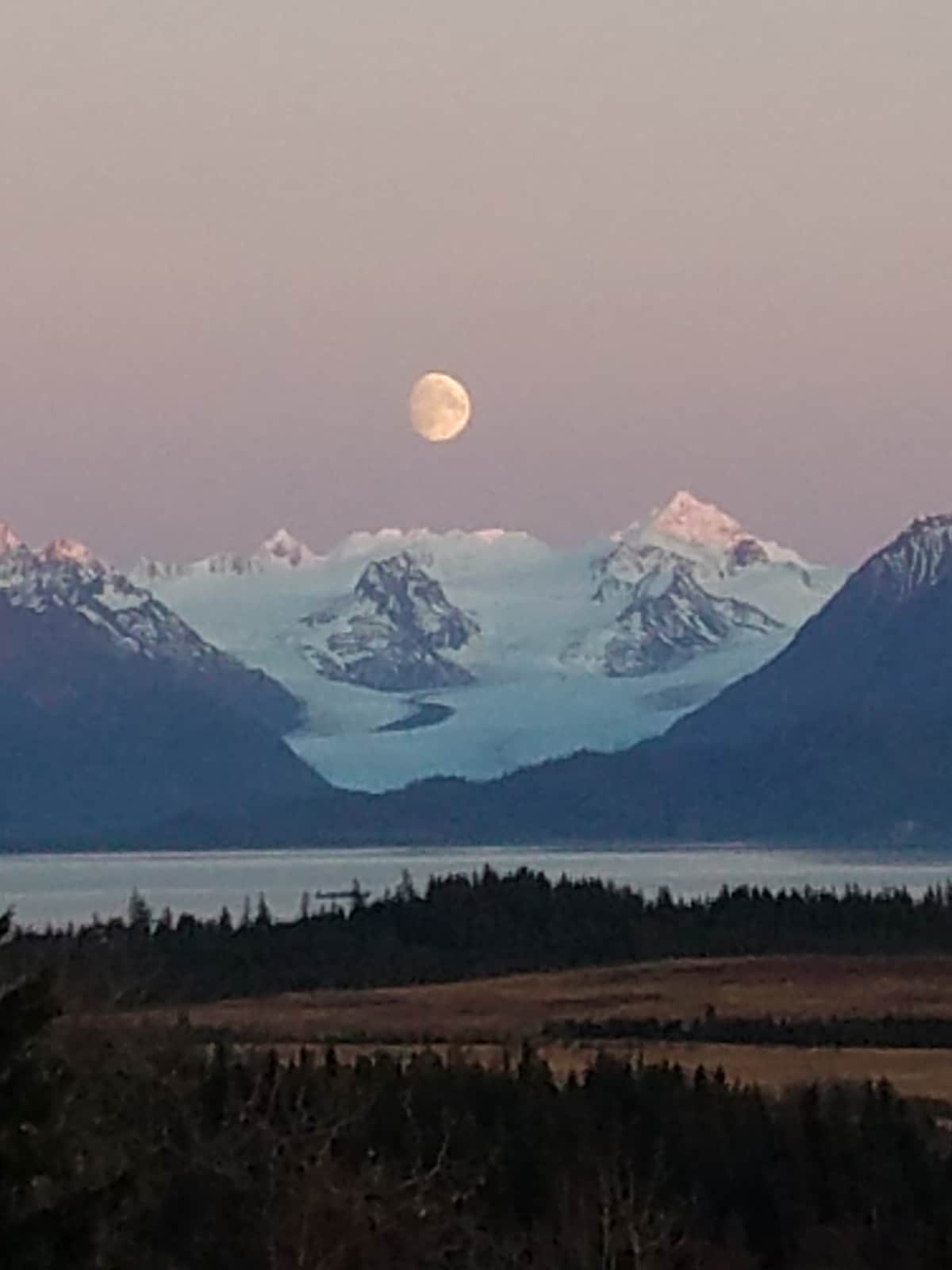A serene landscape reveals majestic mountains topped with snow, with a glacier descending into the valley. A full moon is seen rising above the peaks, casting a soft glow over the scene. The foreground features a mix of dense forest and open meadow.