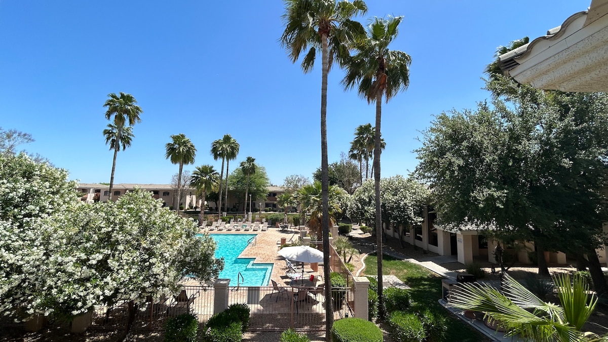 A view of the resort-style pool area is visible, surrounded by palm trees and flowering shrubs. Lounge chairs are arranged near the pool, and an umbrella provides shade. The bright blue sky complements the inviting atmosphere of the outdoor space.